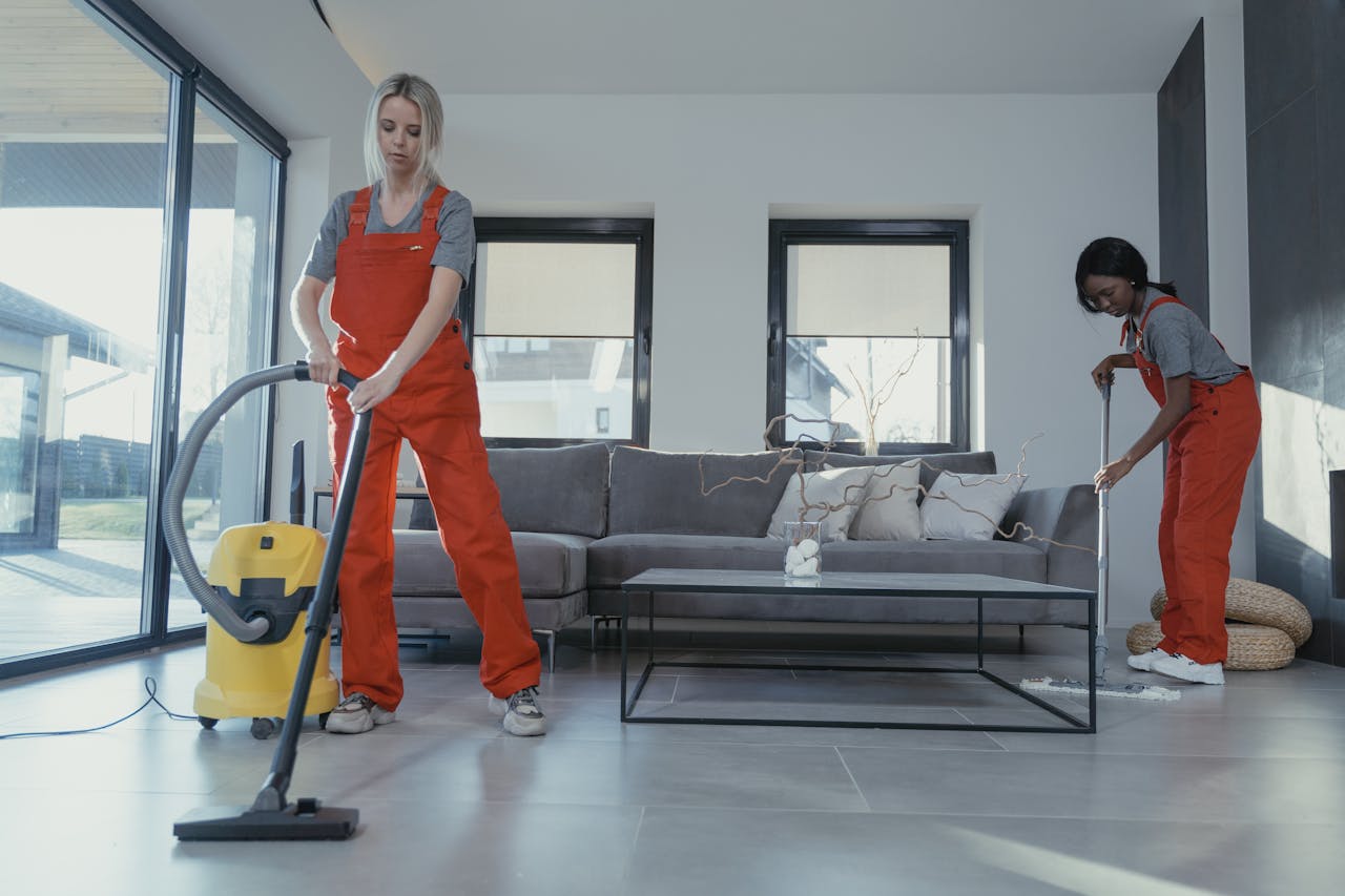 hero-img-01 Two women in red overalls vacuuming and sweeping a modern living room.